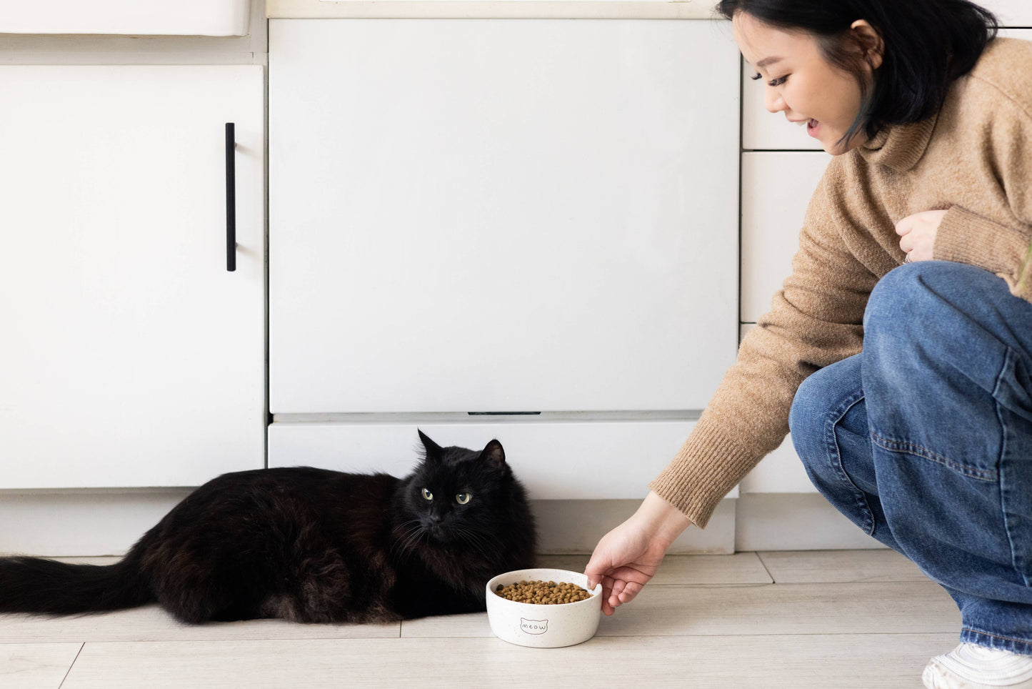 Woman feeding a black cat from a bowl on a kitchen floor.